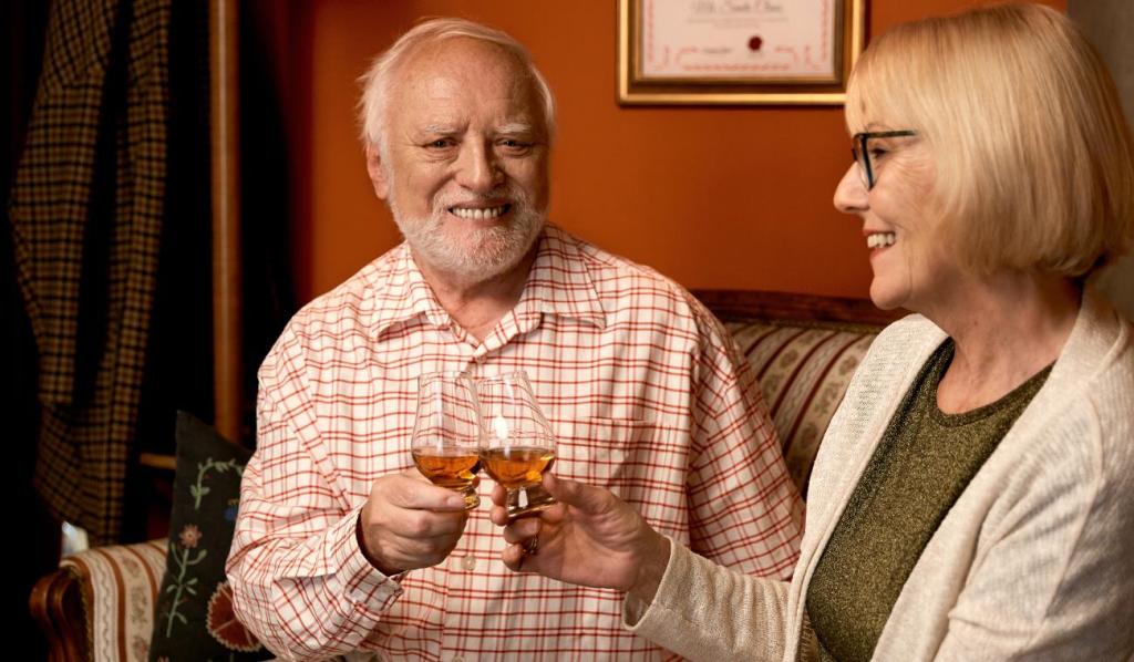 Hide the Pain Harold toasting with a woman with whisky glasses in cozy home interior.